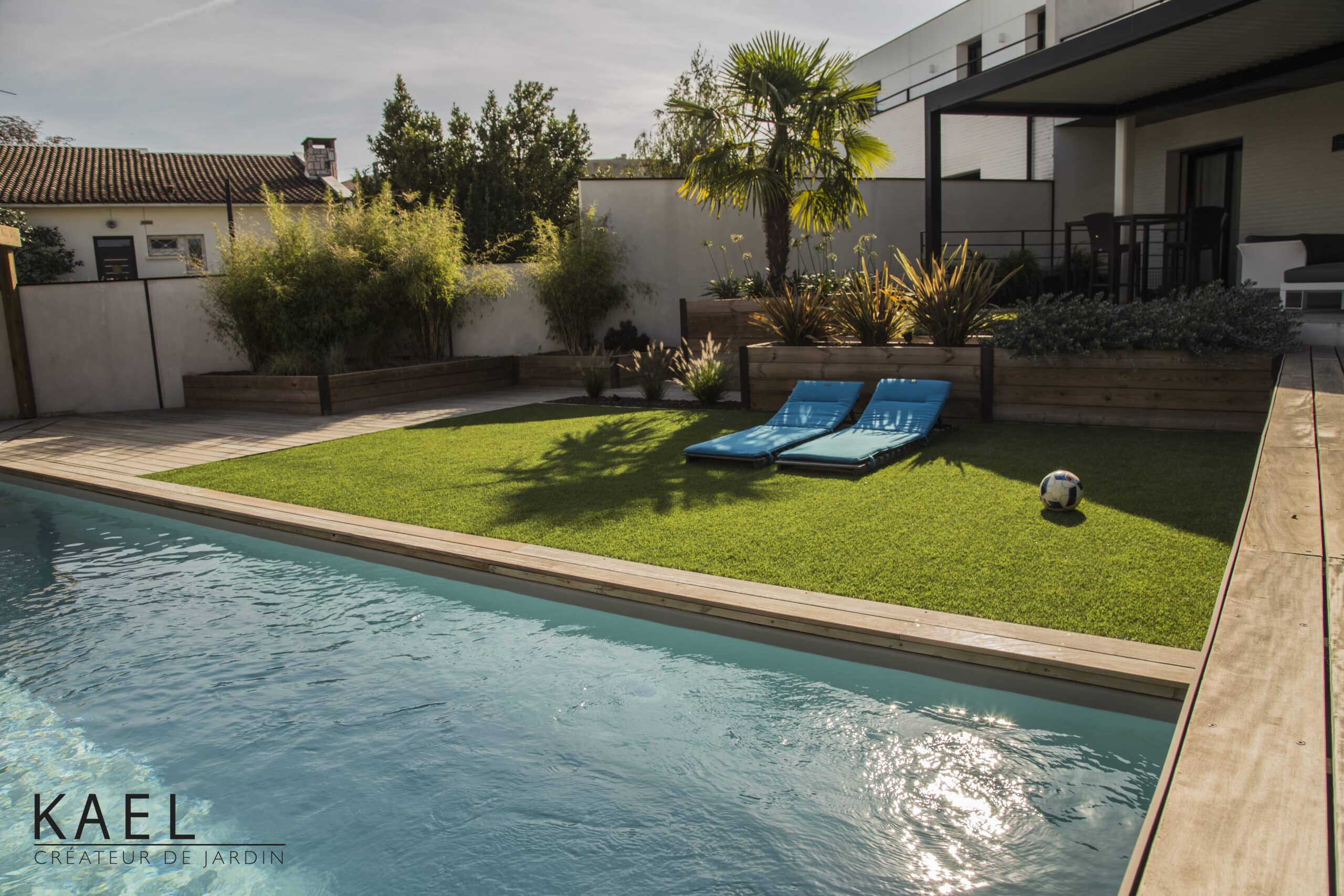 KAEL CRÉATEUR DE JARDIN : piscine, gazon synthétique, transats bleus et palmier dans un jardin design moderne.