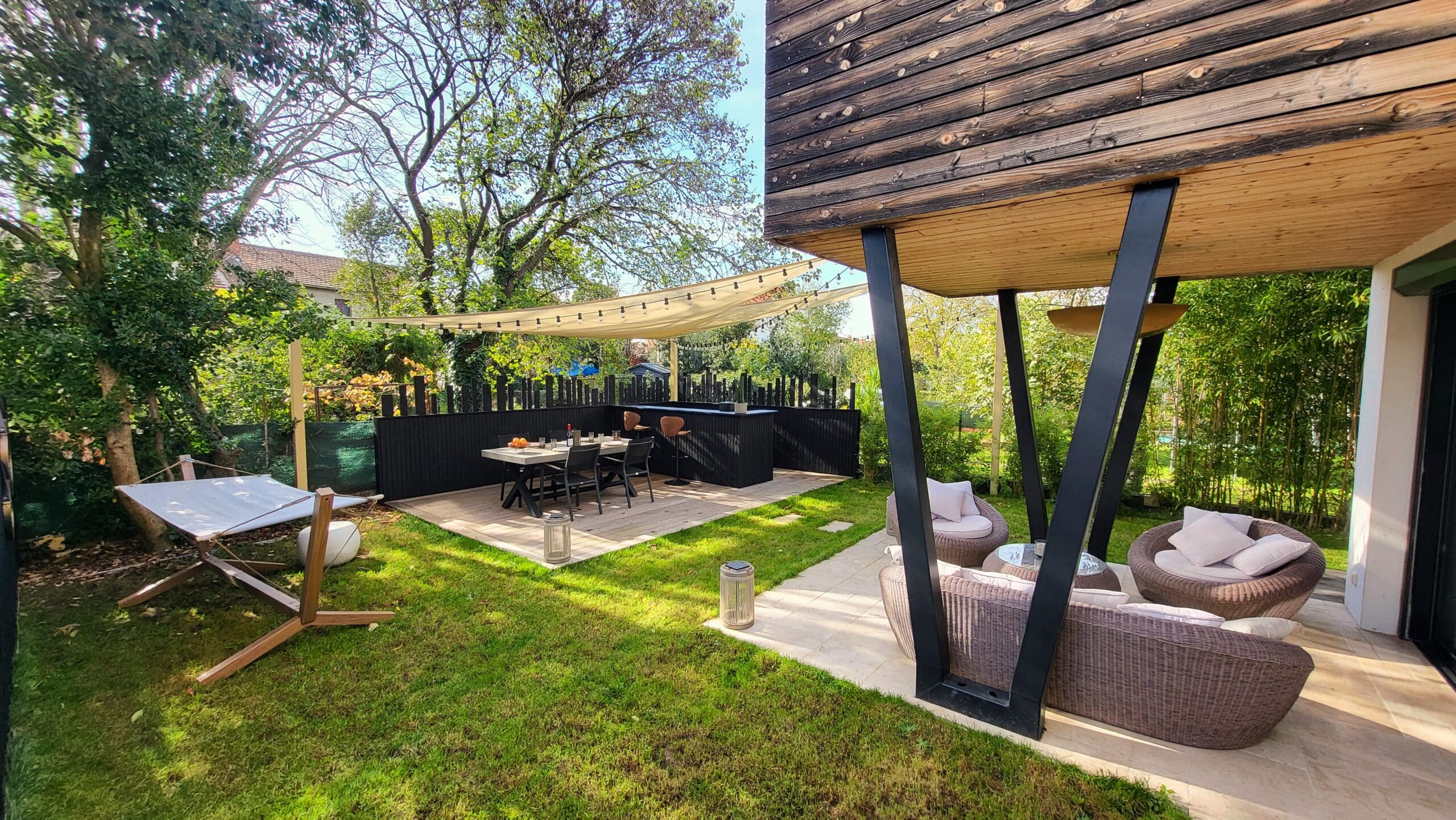 Aménagement jardin moderne : salon lounge sous terrasse en bois, hamac sur gazon et coin repas ombragé avec voile solaire.