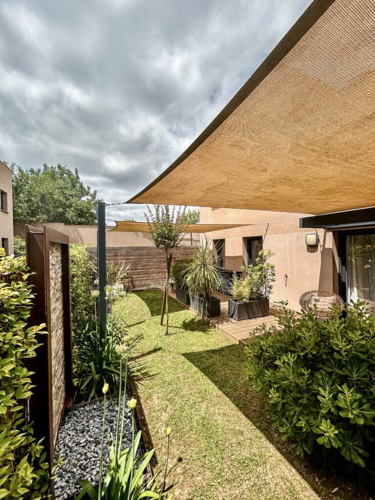 Jardin moderne avec voile d'ombrage beige, terrasse en bois et bacs à plantes sous un ciel nuageux.