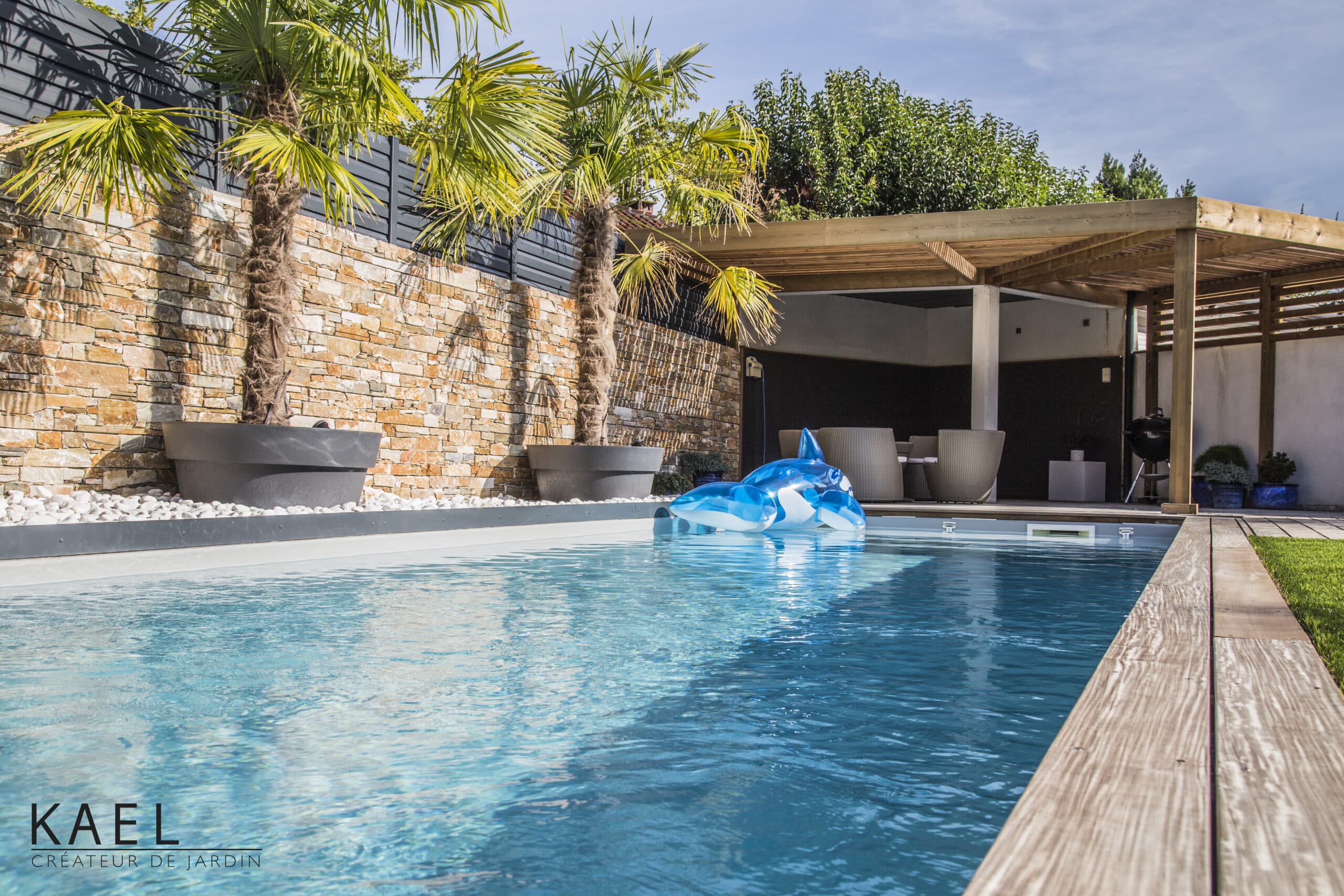 Piscine avec mur en pierre, palmiers en pots et pergola bois. Logo KAEL Créateur de Jardin en bas à gauche.
