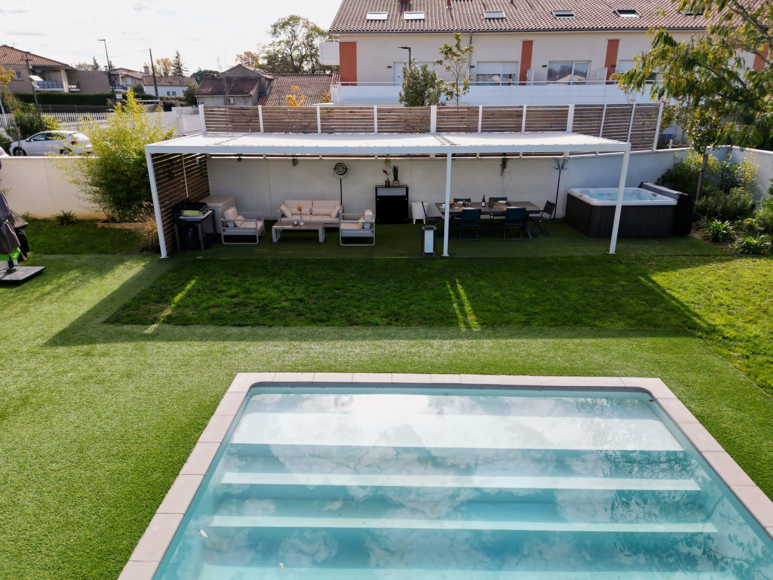 Piscine moderne avec escaliers et pergola blanche abritant un salon de jardin et spa sur gazon synthétique.