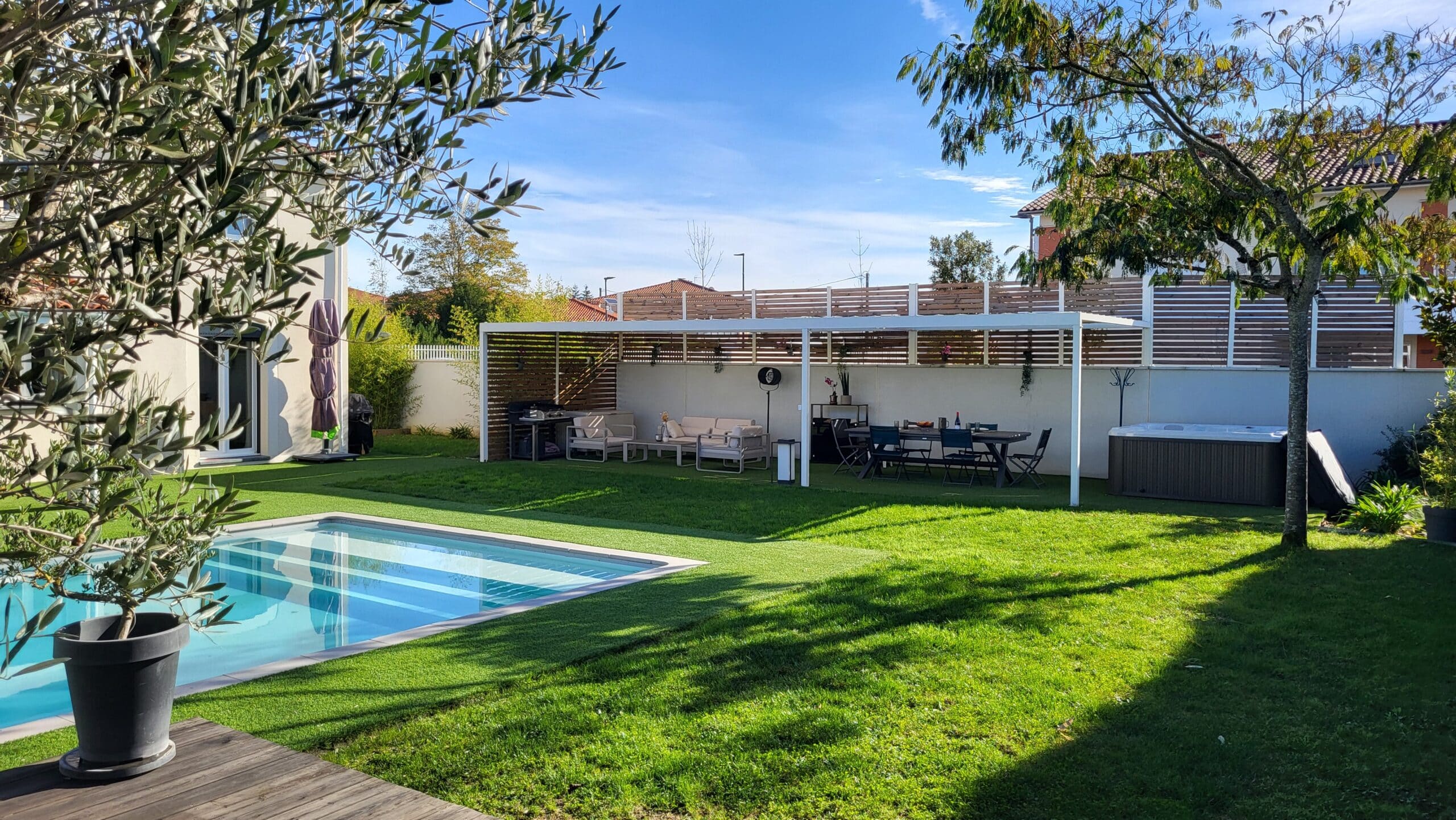 Jardin moderne avec piscine, pergola blanche, salon de jardin, spa et pelouse synthétique par beau temps.