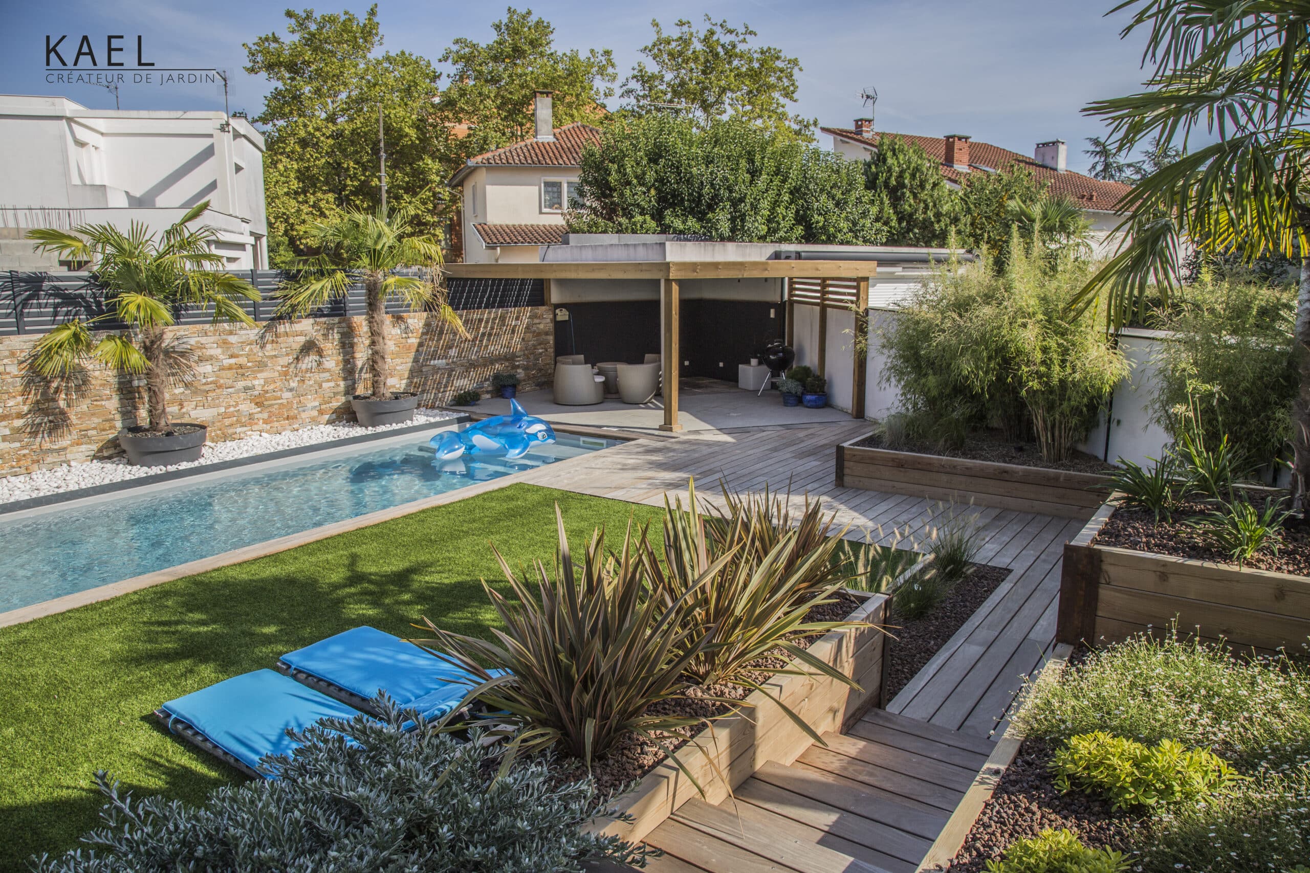 Aménagement paysager KAEL : jardin moderne avec piscine, terrasse en bois, transats bleus et mur en pierre.