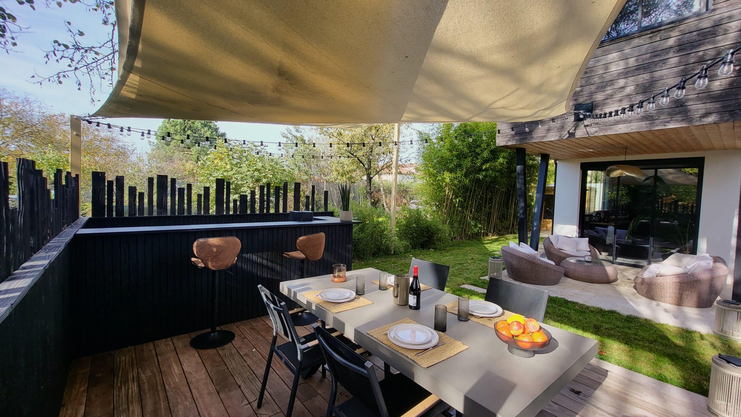 Terrasse moderne avec table à manger, bar noir, voile d'ombrage et salon de jardin dans un cadre verdoyant.
