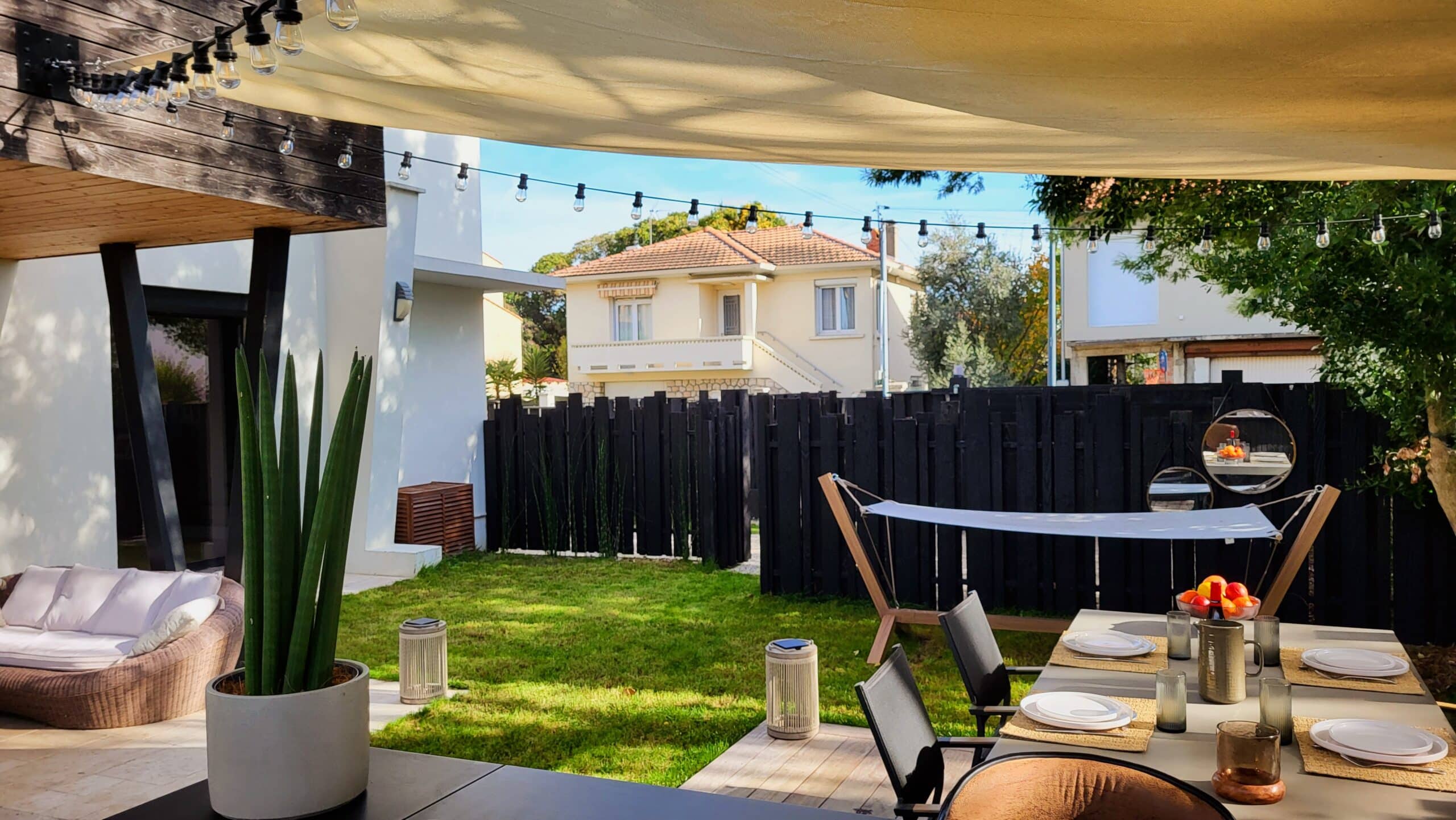 Aménagement terrasse moderne avec table de jardin, hamac et voile d'ombrage sur une pelouse ensoleillée.