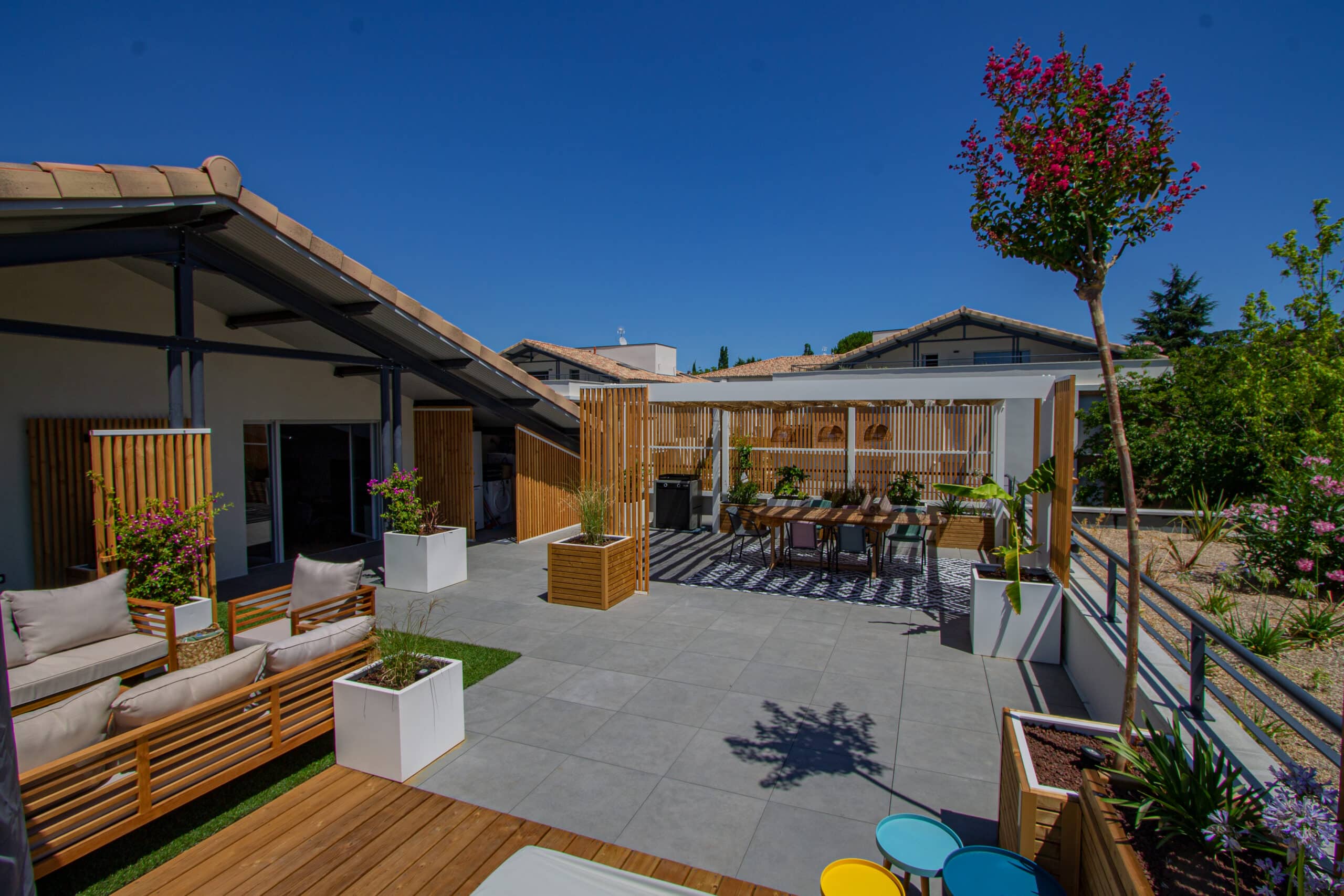 Terrasse moderne aménagée avec pergola, salon de jardin en bois et jardinières fleuries sous un ciel bleu.