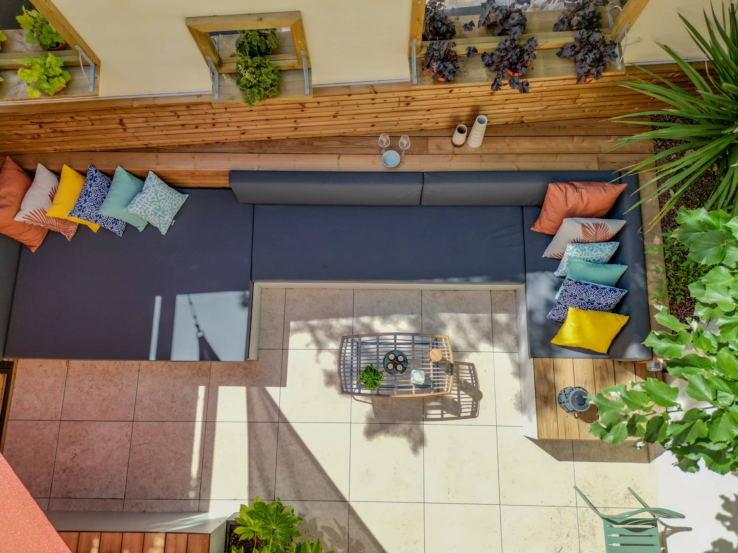Salon de jardin moderne avec canapé d'angle gris et coussins colorés sur une terrasse ensoleillée vue de dessus.