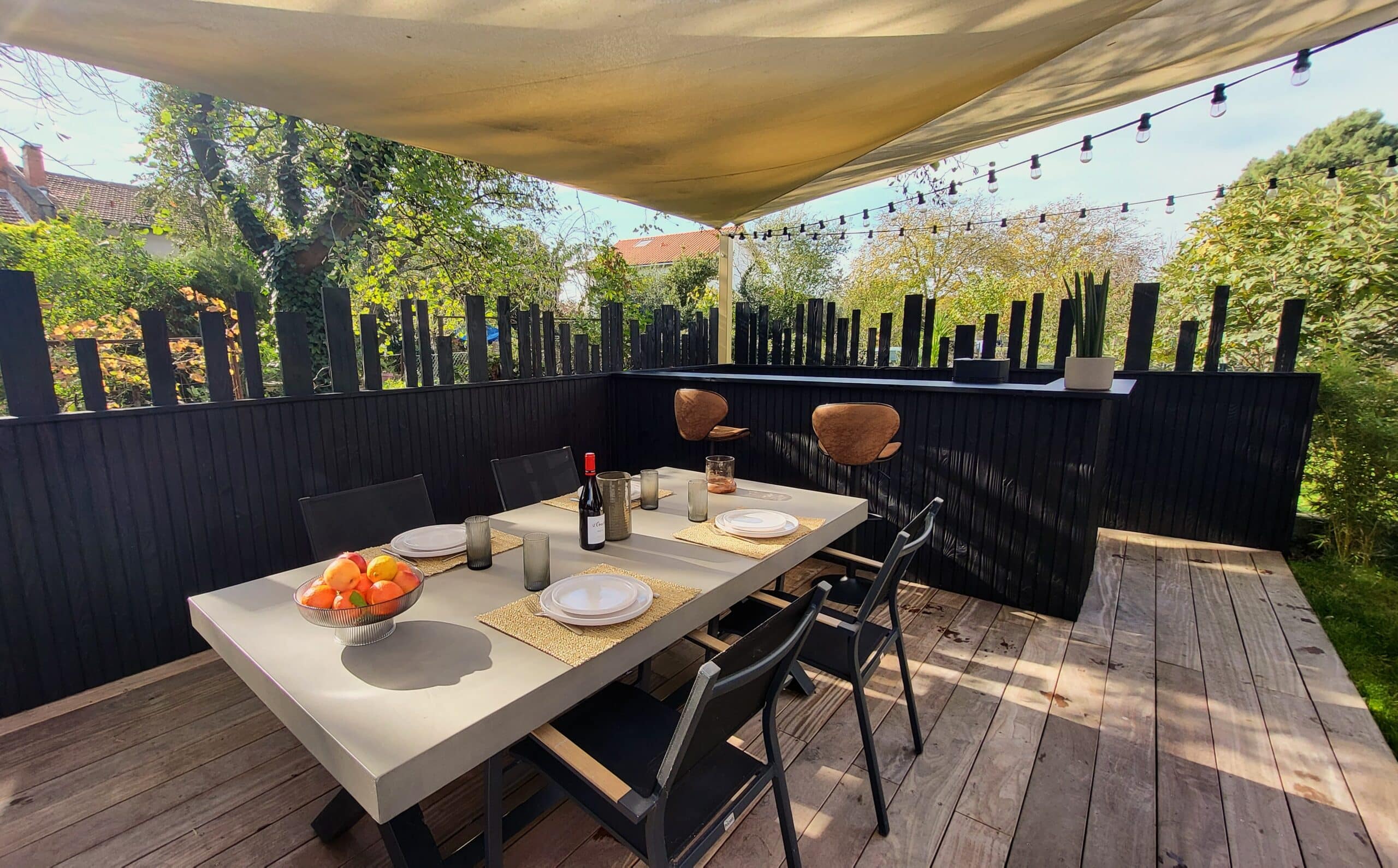 Table à manger grise sur terrasse en bois avec voile d'ombrage, chaises noires et bar extérieur sous des guirlandes.