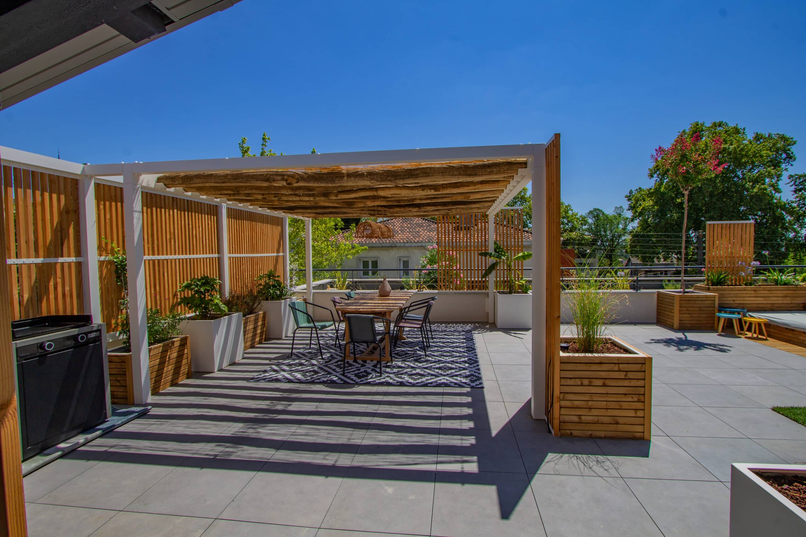 Aménagement de terrasse avec pergola en bois, table à manger et barbecue sous un ciel bleu.