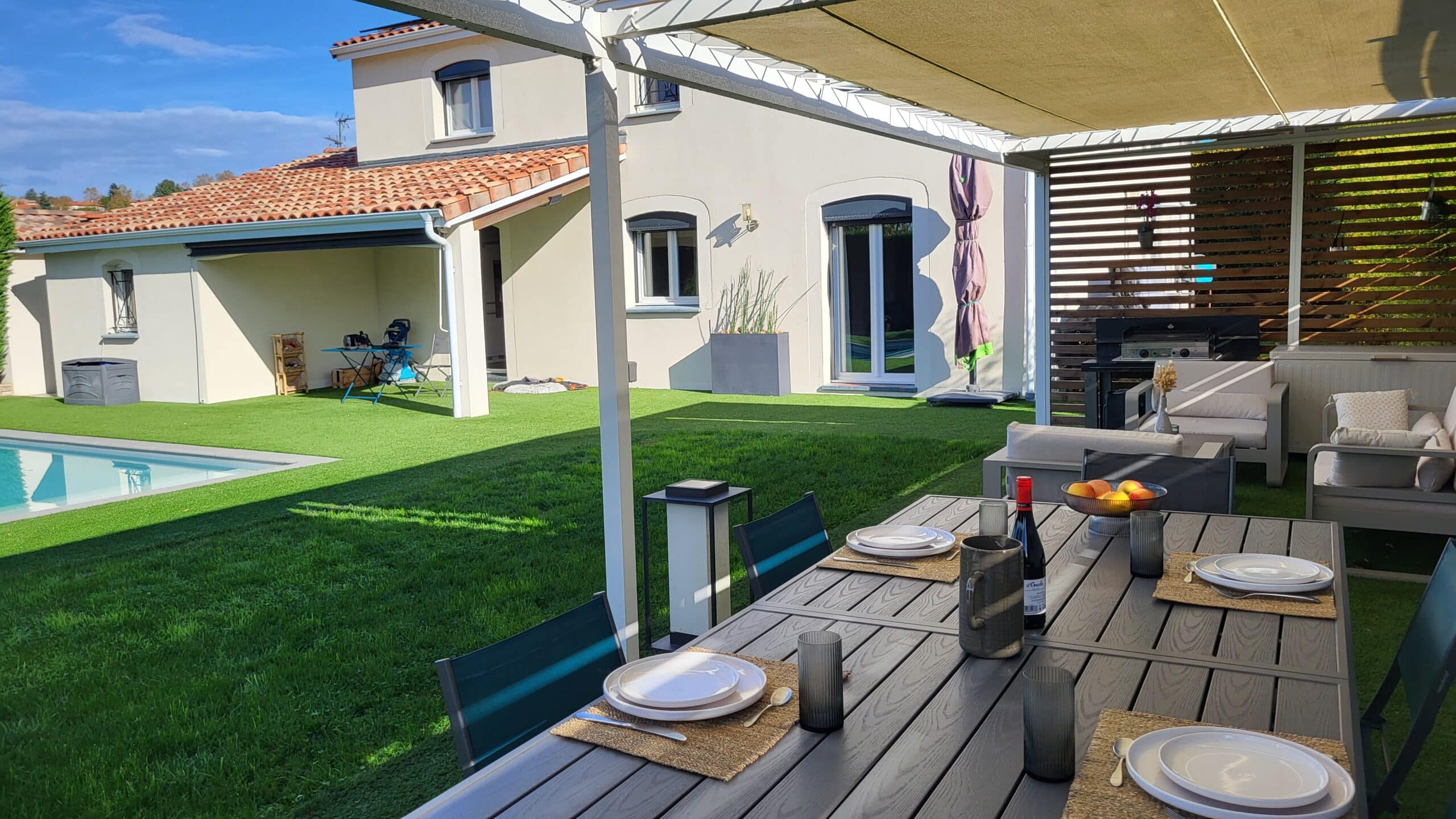 Terrasse avec table dressée sous pergola, vue sur jardin et piscine d'une maison moderne.