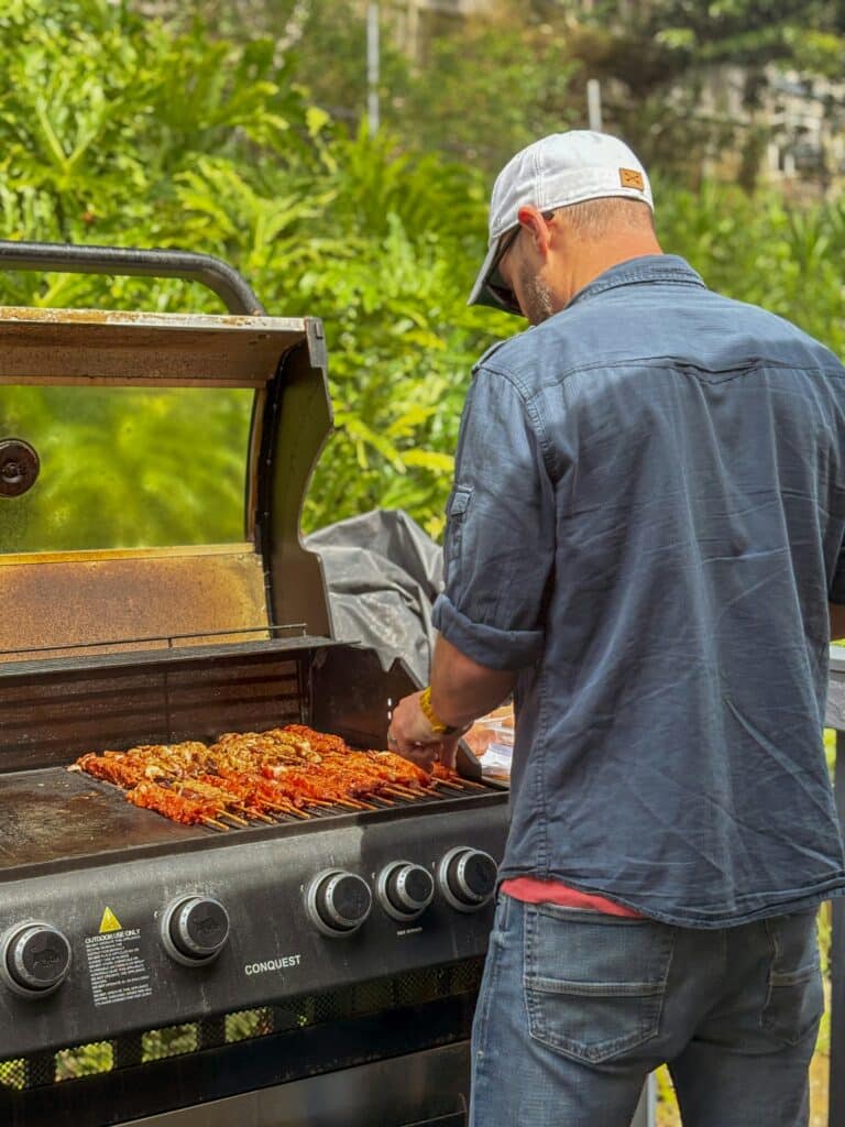 Homme grillant des brochettes de viande sur un barbecue à gaz Conquest devant une végétation luxuriante.