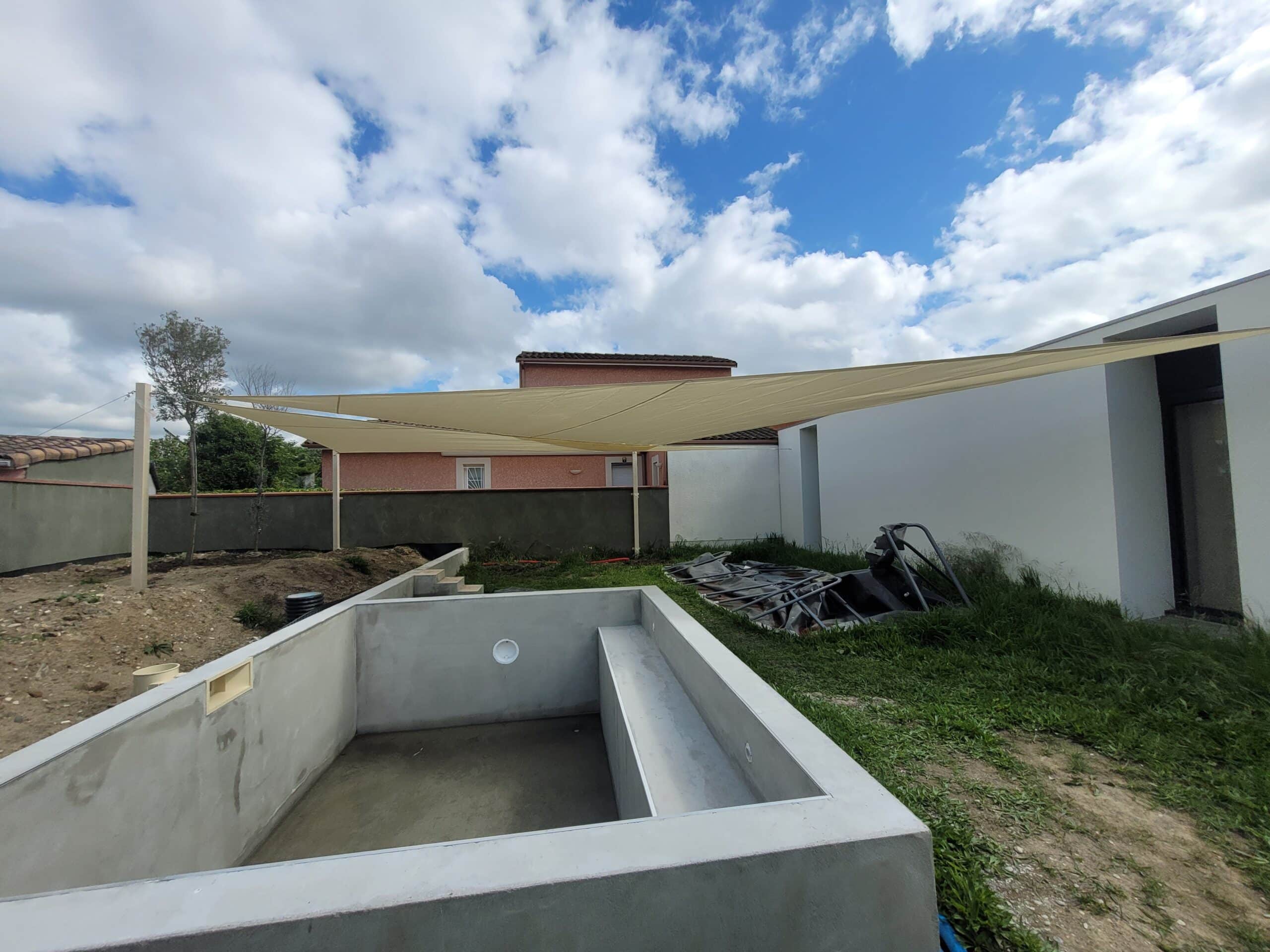 Piscine en béton en construction surmontée d'une voile d'ombrage beige dans un jardin ensoleillé.