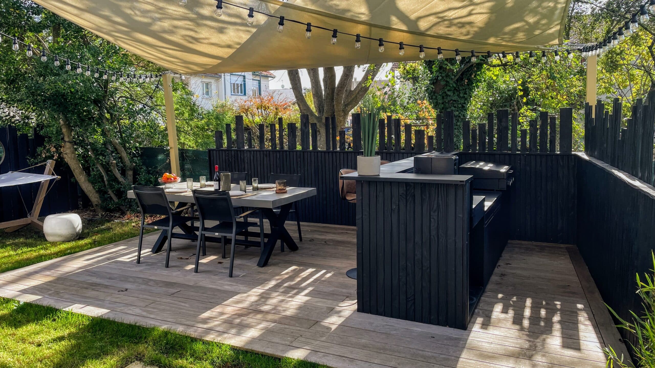 Cuisine d'été et salle à manger moderne sur terrasse en bois avec voile d'ombrage et guirlandes.
