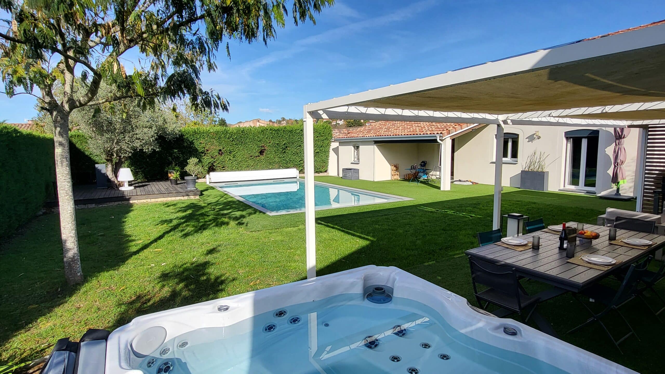 Spa extérieur au premier plan ouvrant sur un jardin avec piscine, pelouse et coin repas abrité par une pergola.