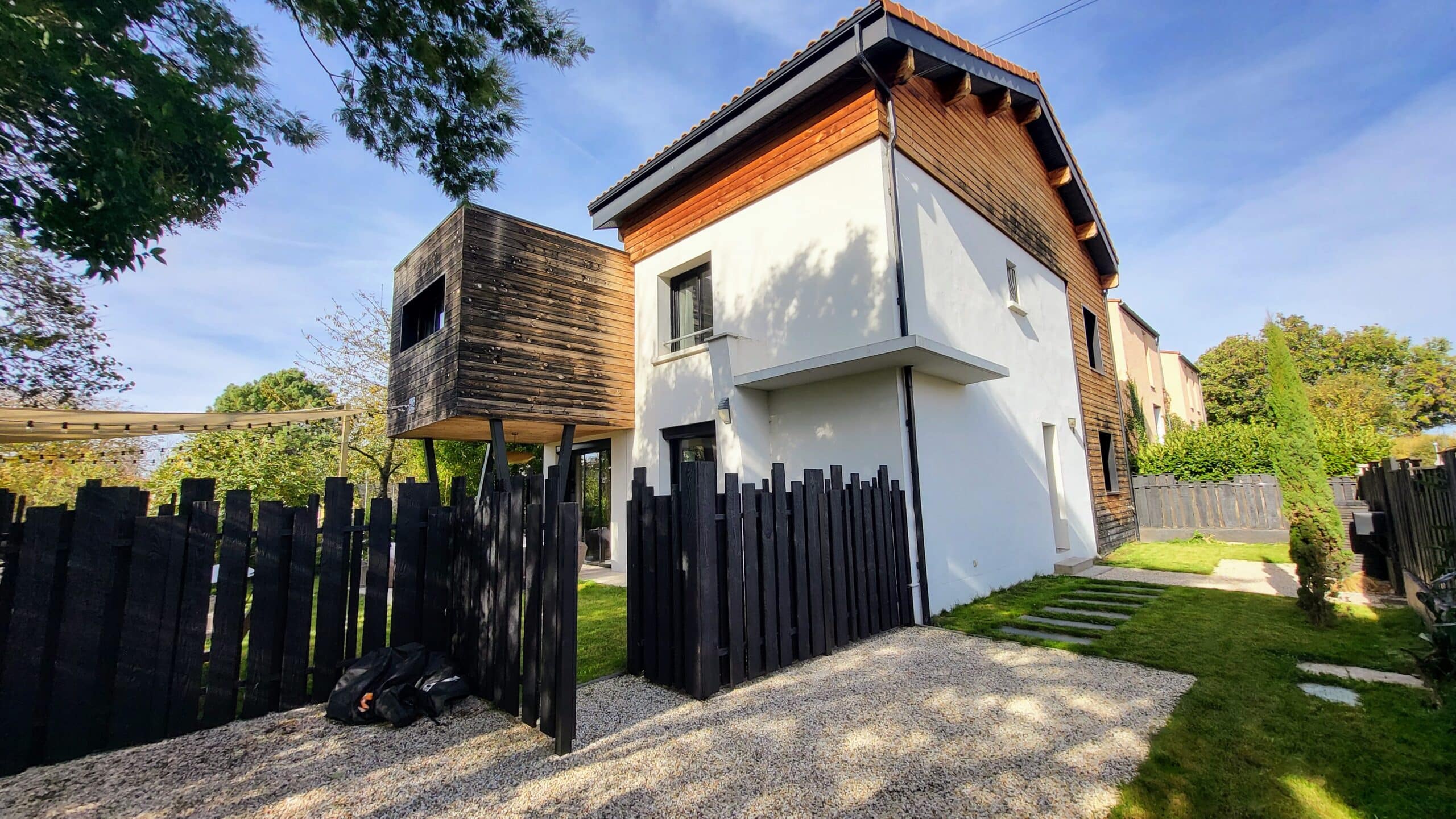 Maison contemporaine avec bardage bois, volume suspendu en bois brûlé, clôture noire et jardin paysager.