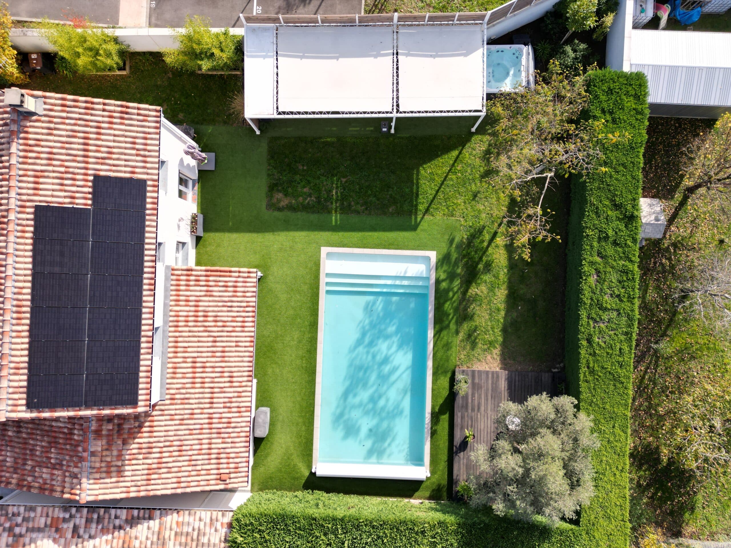 Maison avec piscine, panneaux solaires et pergola vue du ciel au-dessus d'un jardin avec gazon synthétique.