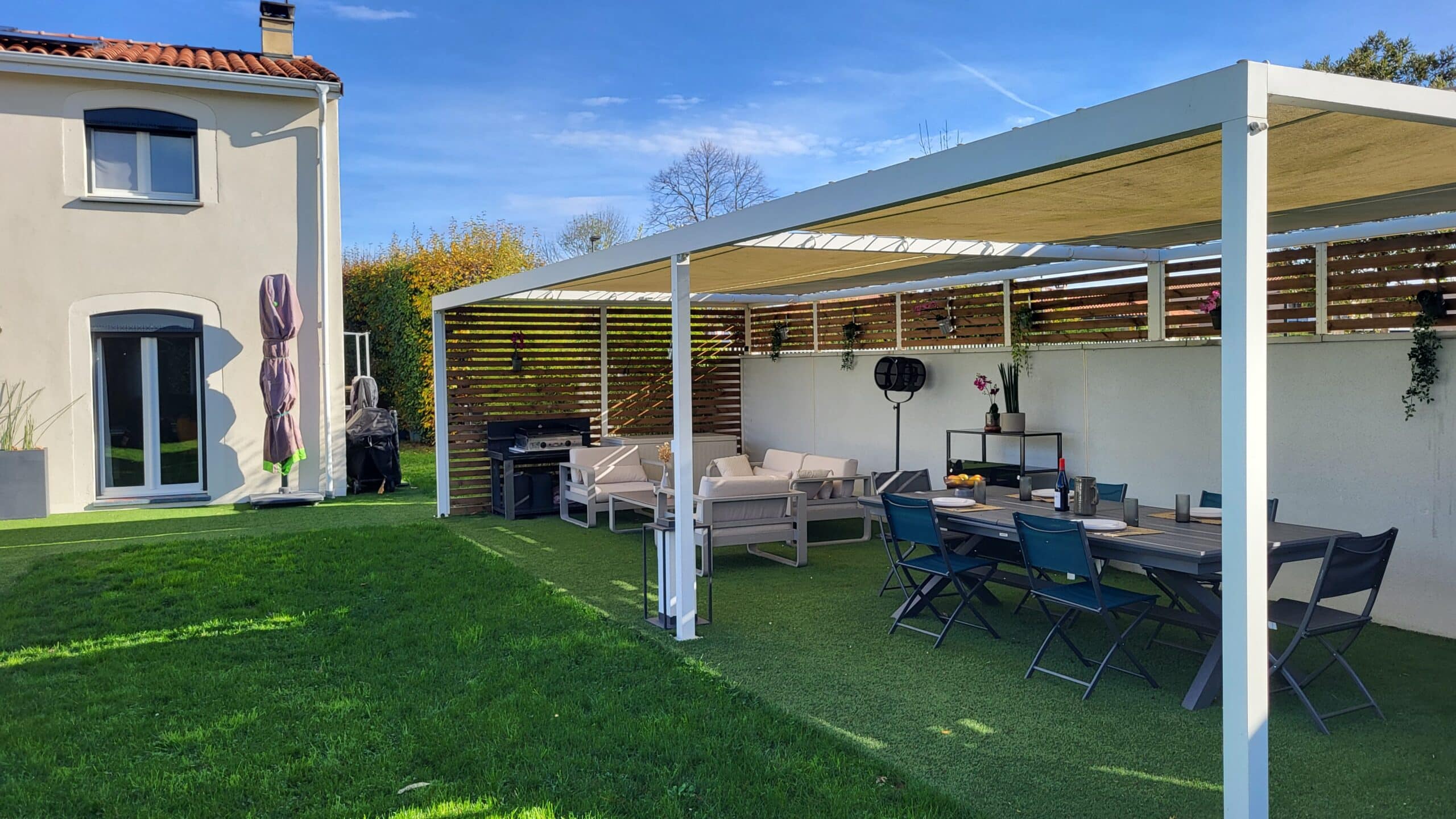Pergola blanche avec salon de jardin, coin repas et barbecue sur gazon synthétique devant une maison moderne.