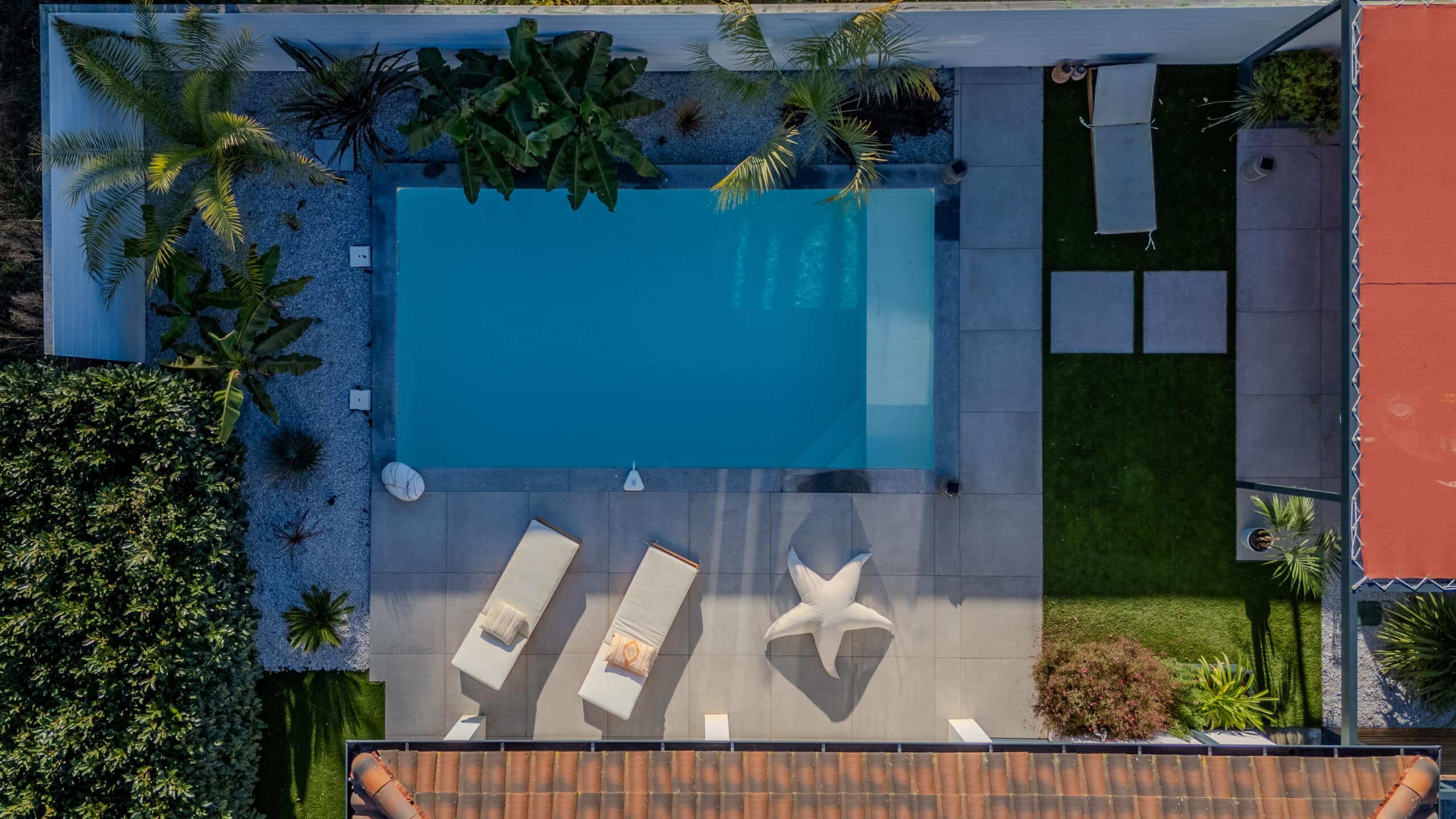Vue aérienne d'une piscine bleue moderne avec chaises longues, pouf étoile et jardin paysager tropical luxueux.