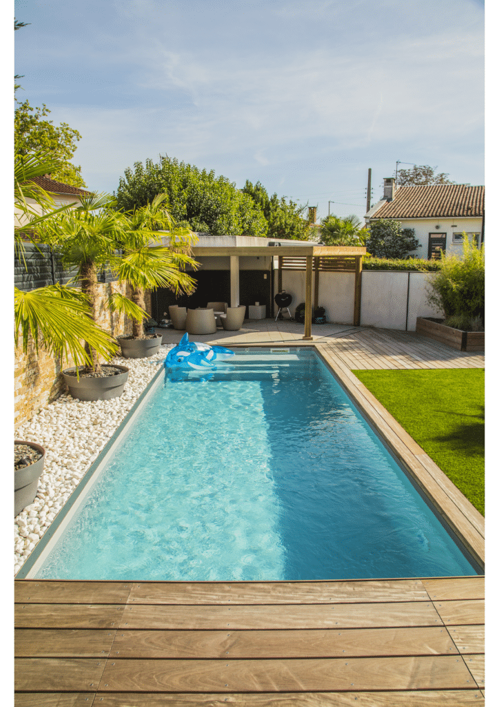 Piscine rectangulaire moderne dans un jardin avec terrasse en bois, palmiers, gazon et bouée baleine bleue.