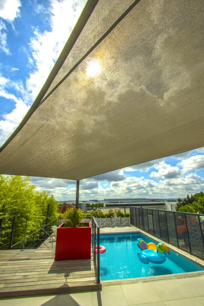 Piscine de toit avec voile d'ombrage, bouée toucan bleue et vue sur la ville sous un ciel ensoleillé.