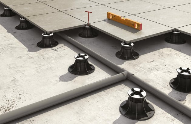 Pose de dalles de terrasse sur plots réglables avec niveau à bulle et canalisations apparentes sur béton.