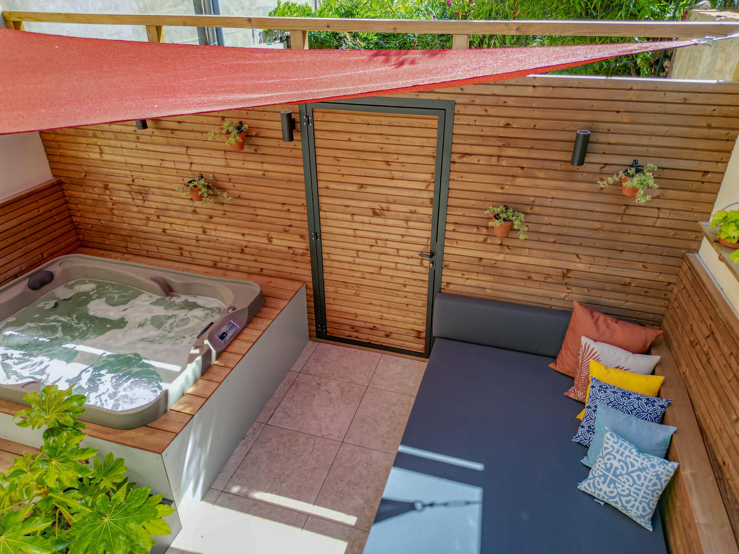 Terrasse détente avec jacuzzi, banquette moderne, voile d'ombrage rouge et murs en bardage bois chaleureux.