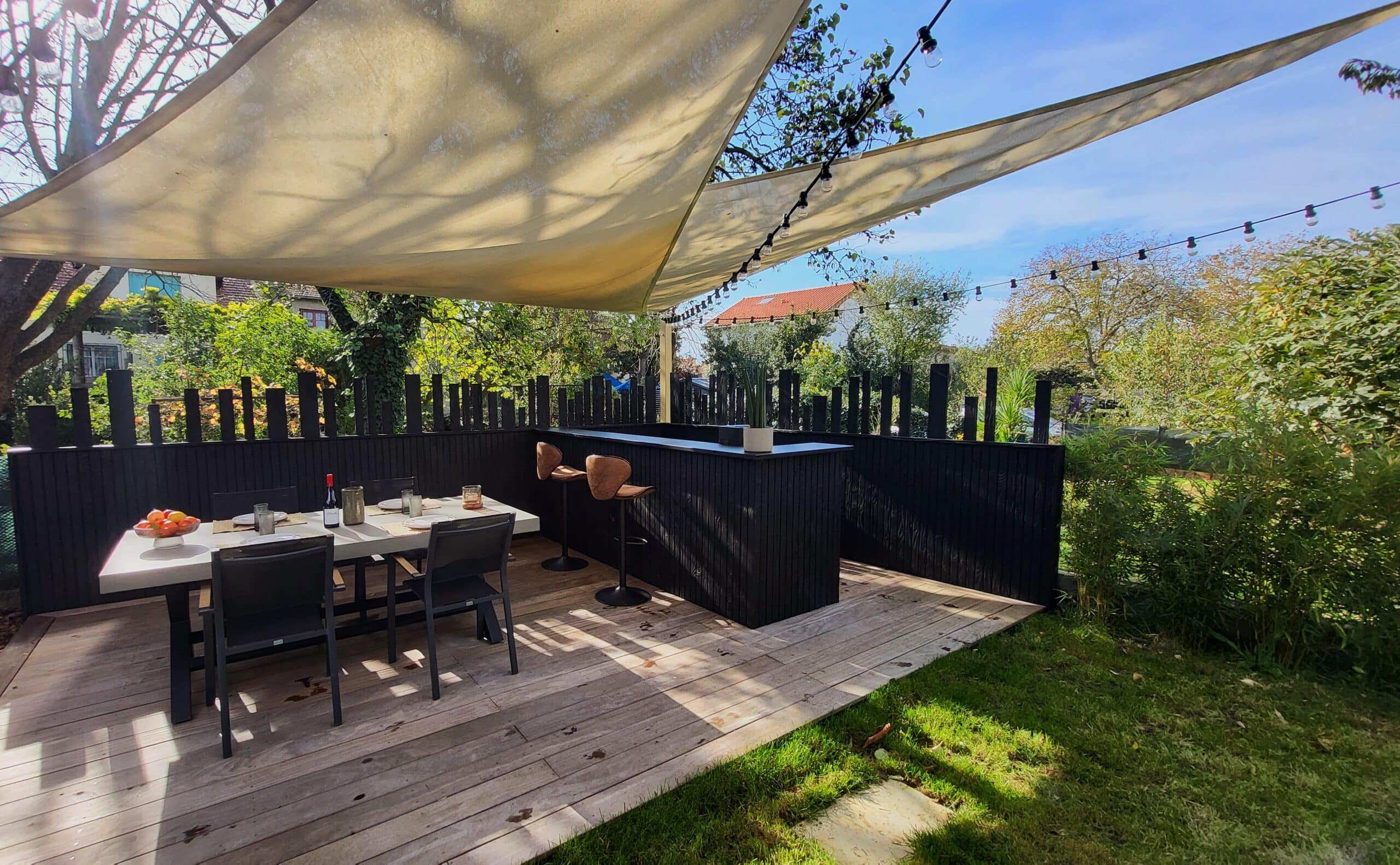Terrasse moderne en bois avec voile d'ombrage, coin repas et bar noir dans un jardin ensoleillé.