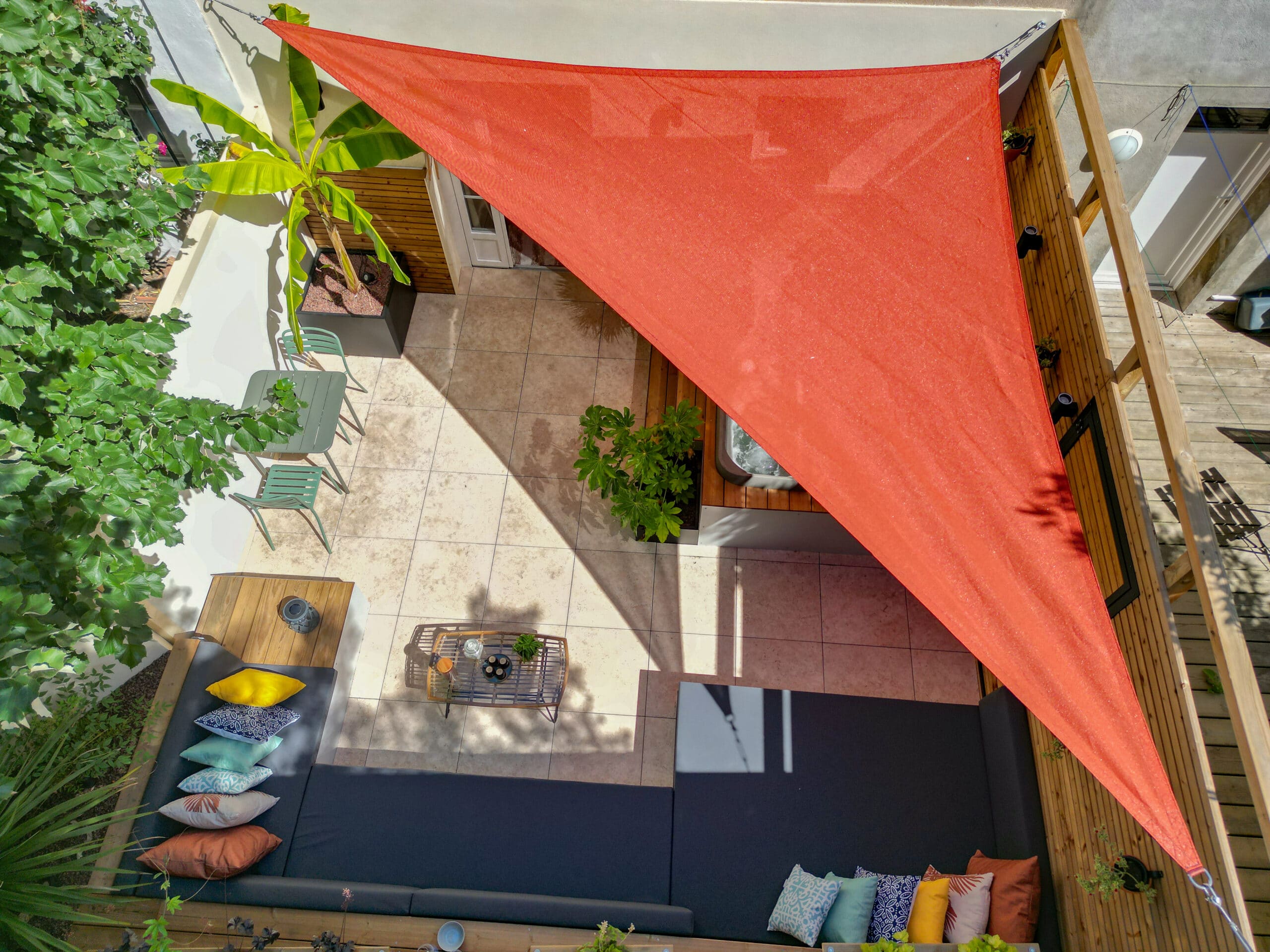 Terrasse moderne avec voile d'ombrage orange, canapé d'angle, table verte, jacuzzi et végétation luxuriante.