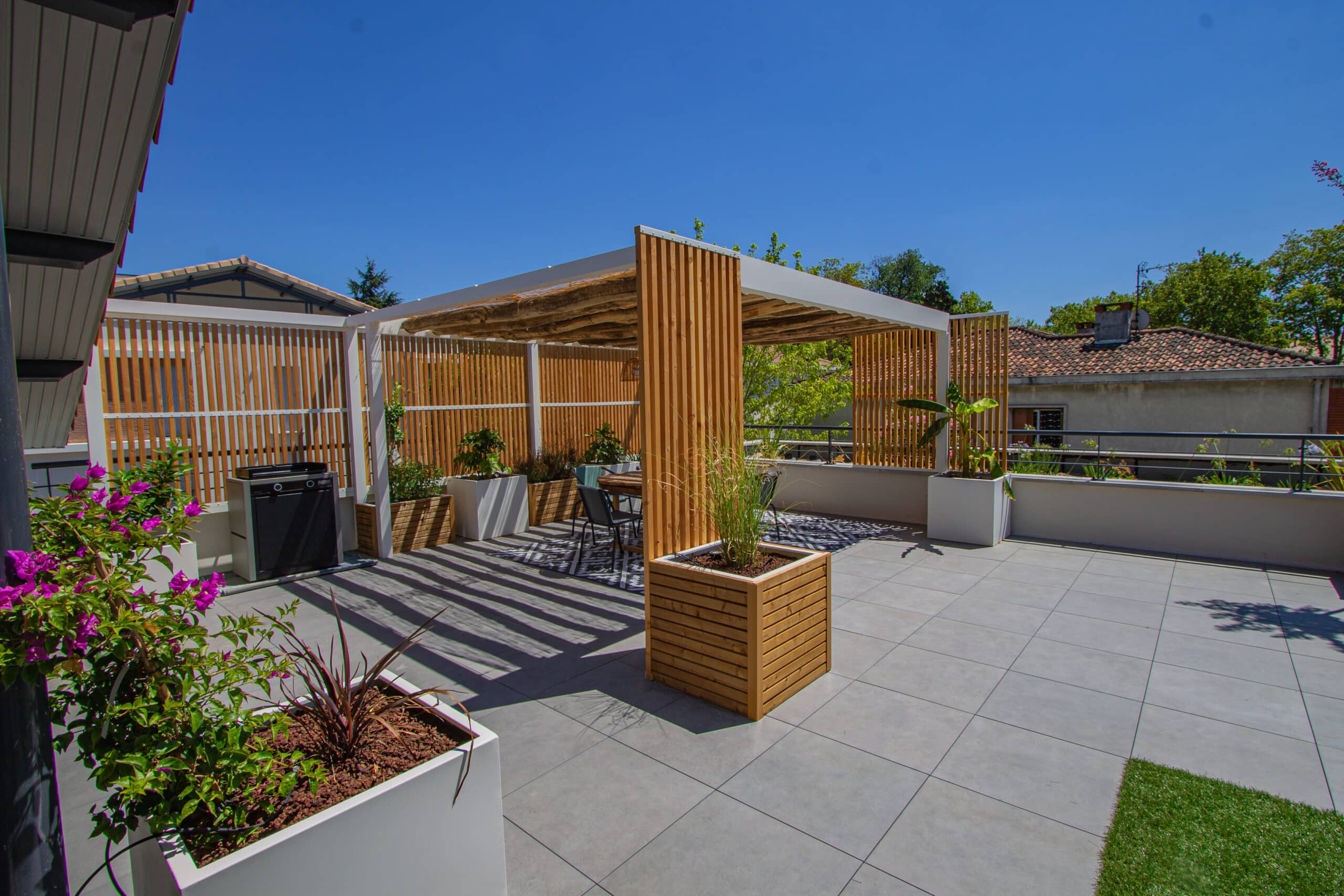 Terrasse de toit avec pergola en bois, jardinières design, carrelage gris et mobilier sous un ciel bleu dégagé.
